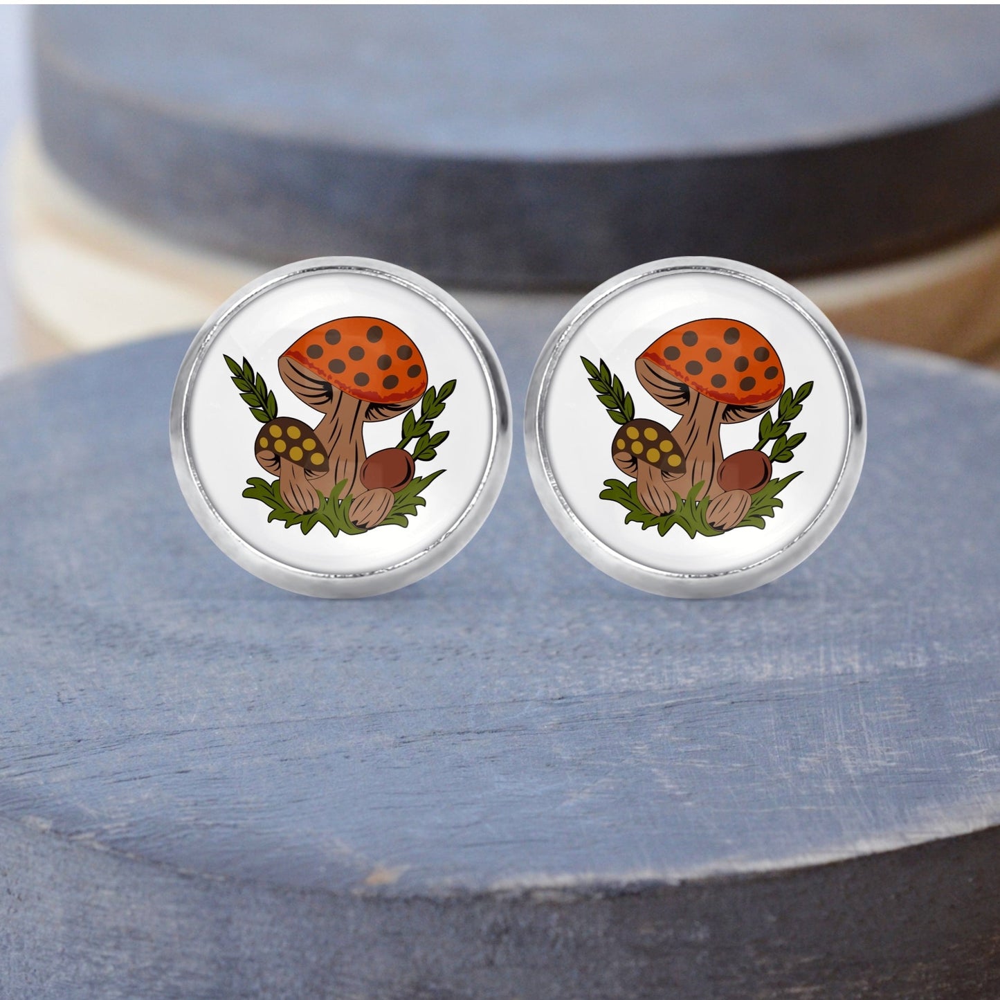 A pair of stud earrings featuring a round Pyrex glass design with a brown and orange Merry mushroom pattern, set in silver-colored post settings, displayed on a gray platform.