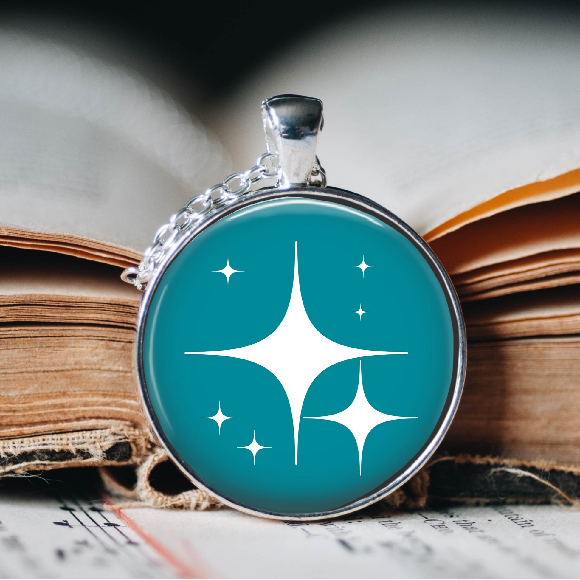 A turquoise and white atomic star pendant necklace with a silver plated chain displayed with a book in the background. 