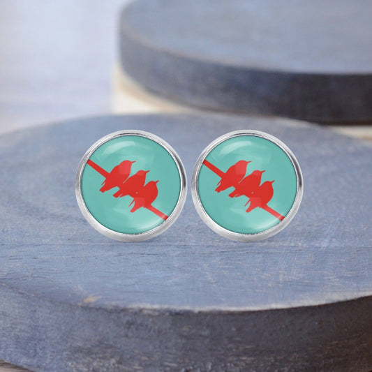 A pair of silver stud earrings with a red bird design on a turquoise background, displayed on a gray stone.